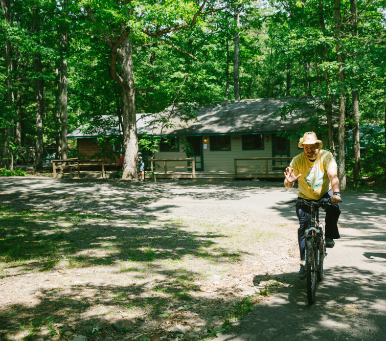 Ben, a counselor at Camp Bonfire, adult summer camp, rides his bicycle through camp.